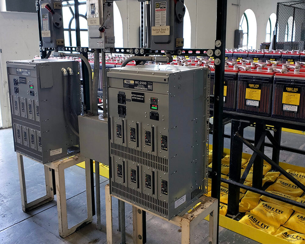 Two Eagle Eye Power Solutions BC-2500 8-bay battery chargers on racks in storage room with racks of batteries behind them.