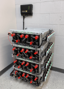 4 short rows of batteries in battery room with wall-mounted monitor and Eagle Eye Power Solutions Vigilant battery monitoring system attached and green sensor lights lit on all sensors
