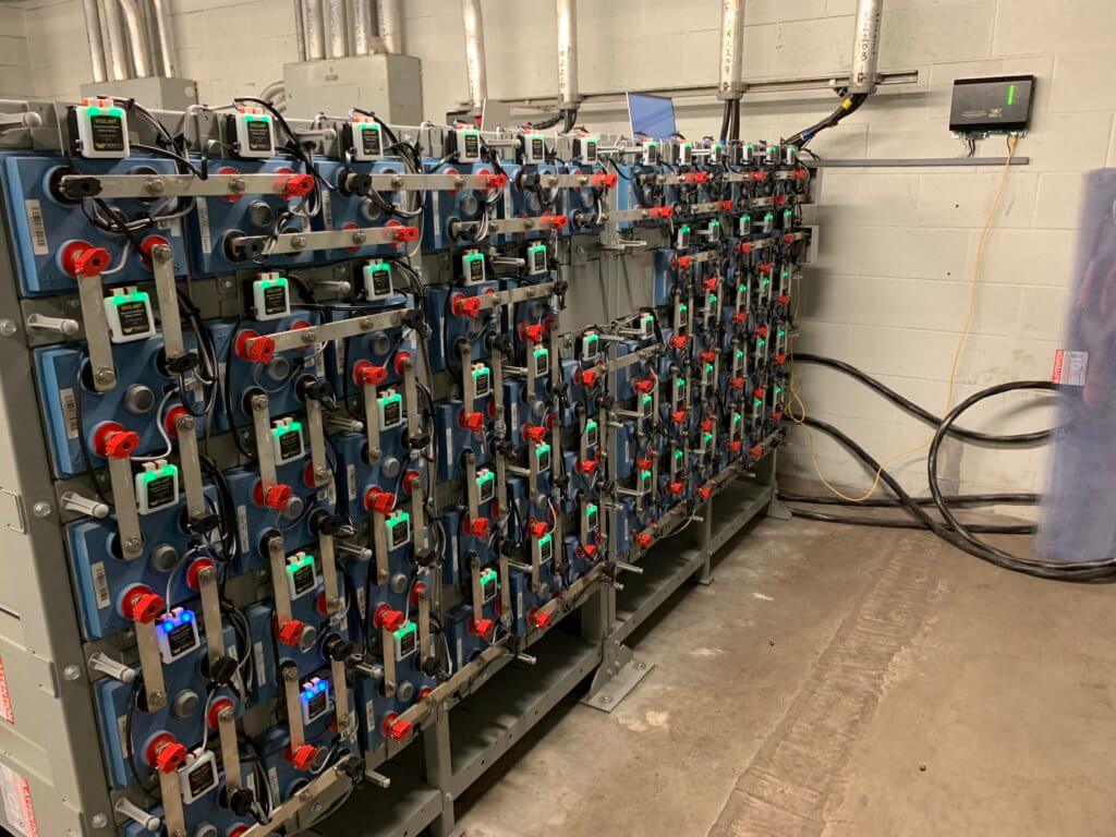 Five racks of batteries in battery room with Eagle Eye Power Solutions Vigilant battery monitoring system attached and sensors displaying green and blue lights, wall-mounted unit displaying green lights.