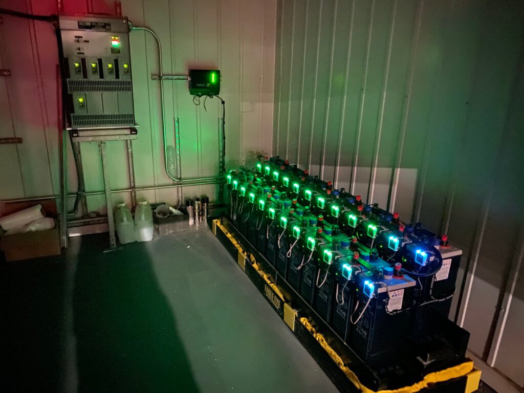 Interior of dark battery room with two rows of batteries with Eagle Eye Power Solutions Vigilant battery monitoring attached and sensors glowing in the dark, green and blue. Green sensors lit up on wall-mounted system.