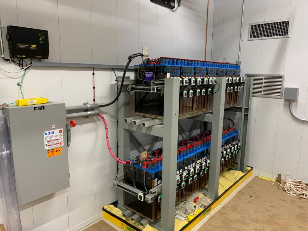 Inside of battery room with two racks of batteries with Eagle Eye Power Solutions Vigilant battery monitoring system attached, green sensors attached, wall-mounts