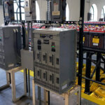 Two Eagle Eye Power Solutions BC-2500 8-bay battery chargers on racks in storage room with racks of batteries behind them.