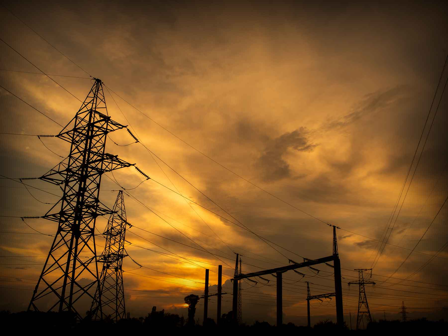 High-voltage power transmission towers silhouetted against a dramatic golden sunset, with a network of cables stretching across the horizon, symbolizing energy distribution and infrastructure.