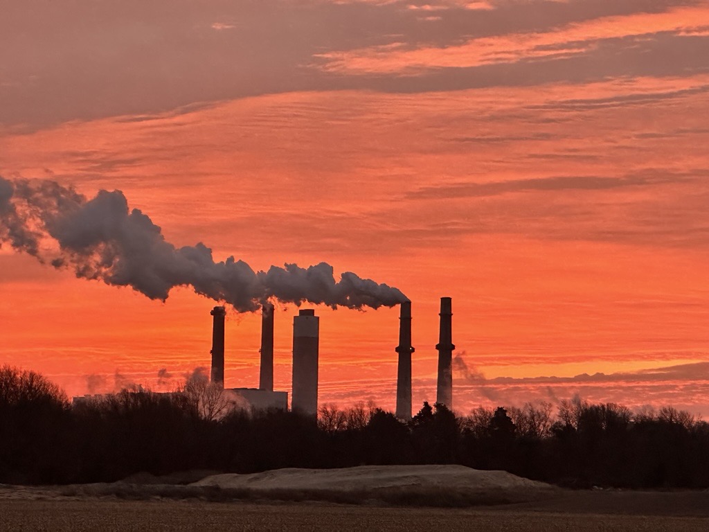 A fiery orange sunrise silhouettes the NIPSCO power plant with four smokestacks, surrounded by trees and a flat landscape.
