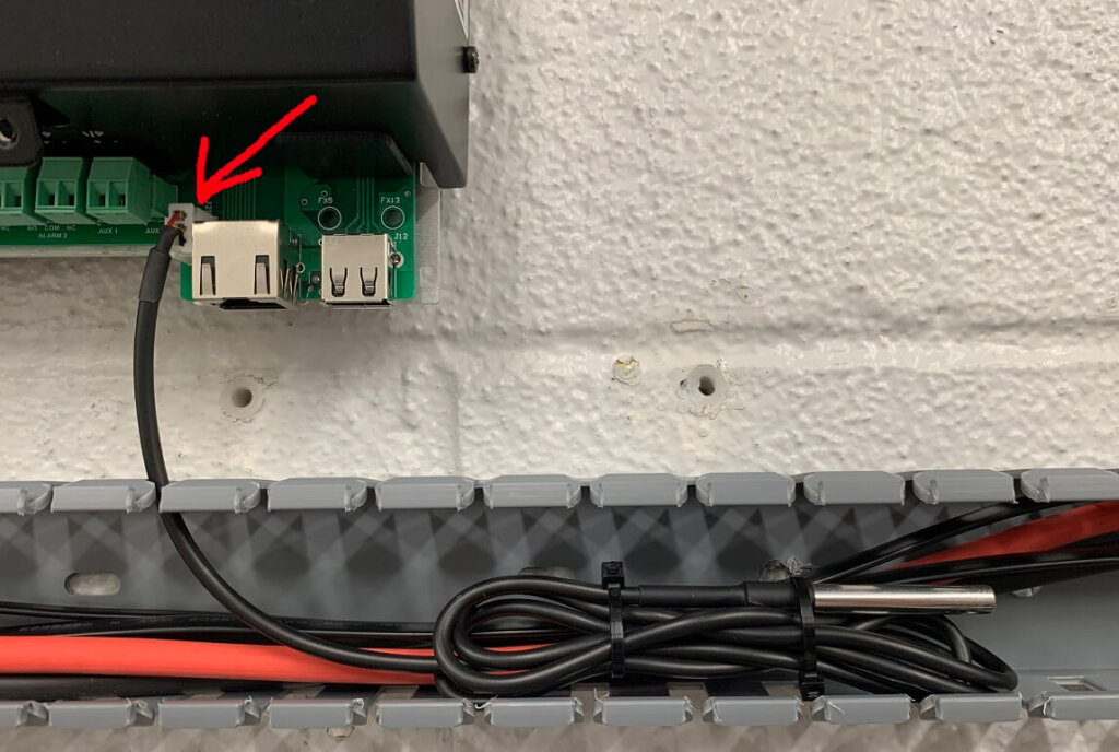 Image showing the ambient temperature sensor plugged into the designated port on the Vigilant® battery monitor circuit board. A red arrow points to the black sensor cable connected to the board near two USB ports, with the sensor itself secured neatly in wire ducting below.