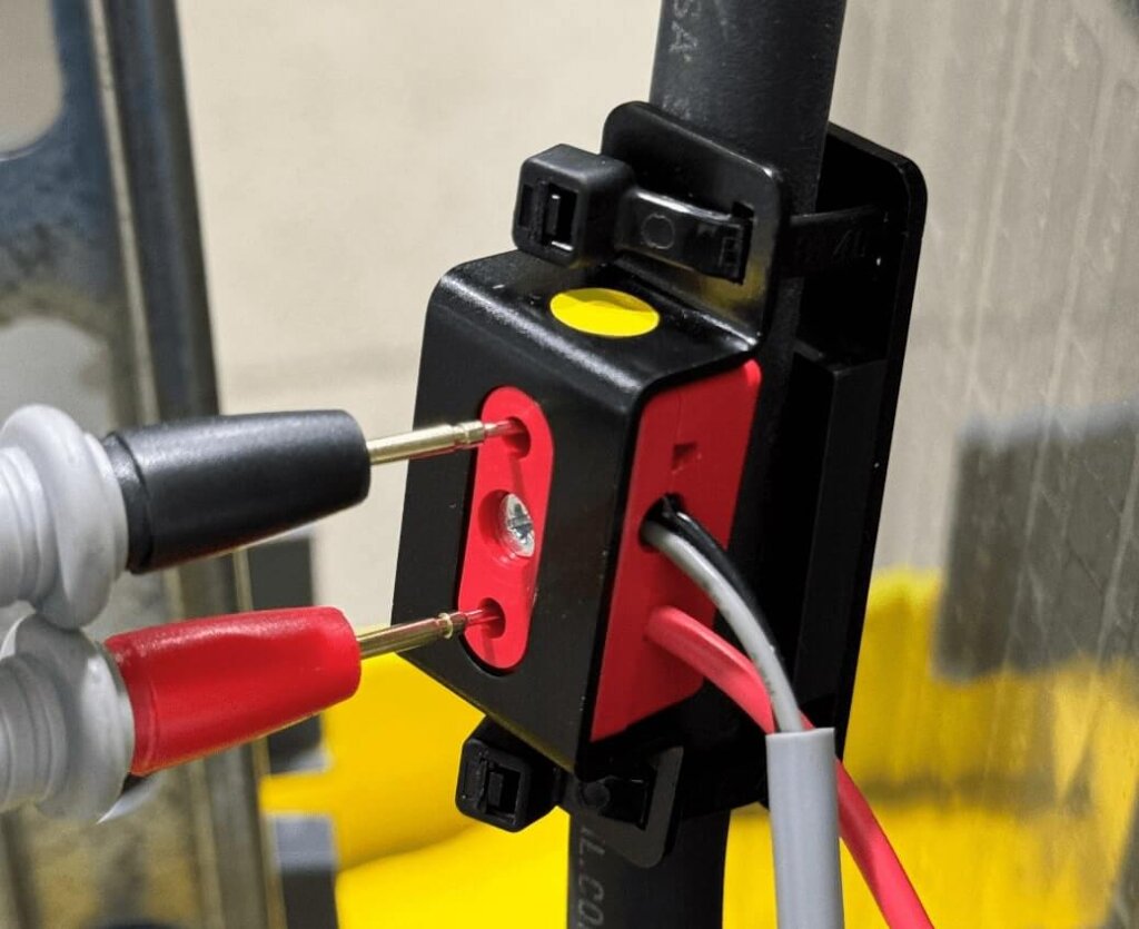 Close-up image of a battery sensor being tested with two probe leads inserted into red test points. The sensor is clamped onto a black battery cable, with red and gray wires connected to its side. The test setup is secured in place using a mounting bracket, and the background shows part of a battery system with yellow insulation.