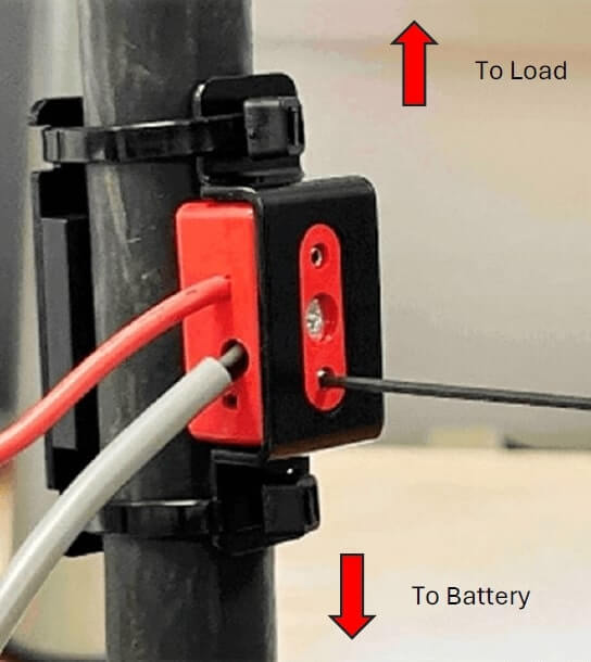 A red and black clamp is secured around a charger cable, with two wires (gray and red) exiting the left side. A red arrow labeled "To Load" points upward, indicating the correct orientation of the clamp, while a red arrow labeled "To Battery" points downward, showing the cable direction back to the battery.