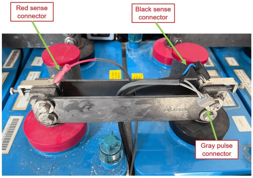 Battery connection setup showing proper placement of Vigilant® connectors. The red sense connector is attached to a clamp on the positive post, the black sense connector is attached to a clamp on the negative post, and the gray pulse connector is connected to a tab washer on the negative post. Each connector is clearly labeled for identification.