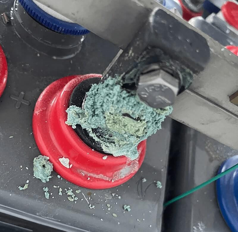 Close-up image of a heavily corroded battery terminal with a buildup of blue-green corrosion on the post and connector hardware. The image highlights the need for cleaning battery terminals and removing excess corrosion or grease before installing Vigilant® battery monitoring equipment to ensure accurate resistance measurements and secure mounting.