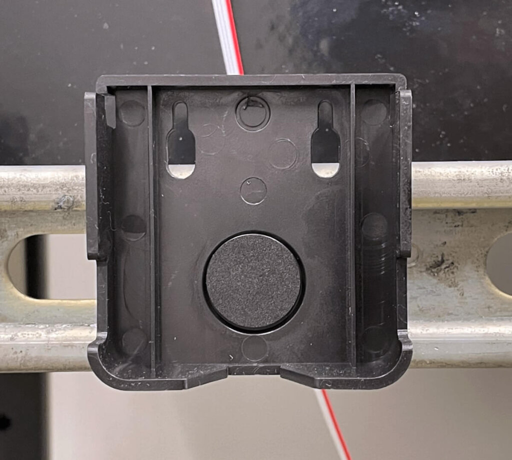 Close-up of a cradle mount for an Eagle Eye Power Solutions ELM sensor attached to a DIN rail. The mount is ready for sensor installation, with a black dot sticker covering the central screw, following the instruction to conceal mounting hardware after securing the cradle to a surface.