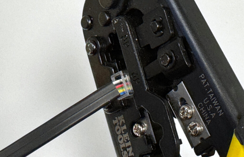Close-up image of a Klein Tools crimping tool being used to crimp an RJ12 connector attached to a black flat cable. The connector is properly inserted into the "6P" slot on the tool, and the cable wires are aligned inside the connector, ready for crimping.