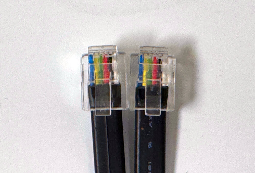 Close-up of two RJ12 connectors placed side by side, showing the correct wire orientation for a crimped cable. Both connectors display the same color sequence from left to right—blue, yellow, green, red, black, and white—emphasizing proper alignment to prevent sensor or monitor damage.
