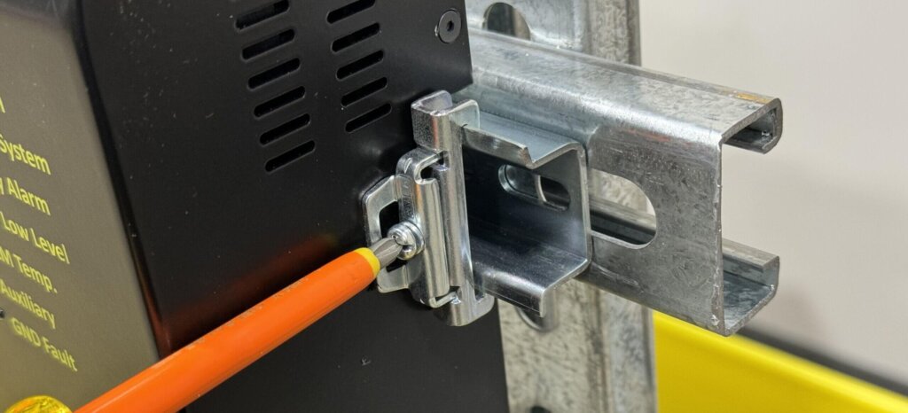A close-up image of a DIN rail stop being installed on the side of an Eagle Eye Power Solutions monitor. An insulated screwdriver is used to tighten the stop, which is positioned on a metal DIN rail. The stop is shown properly inserted and gripping both edges of the rail to secure the monitor in place.