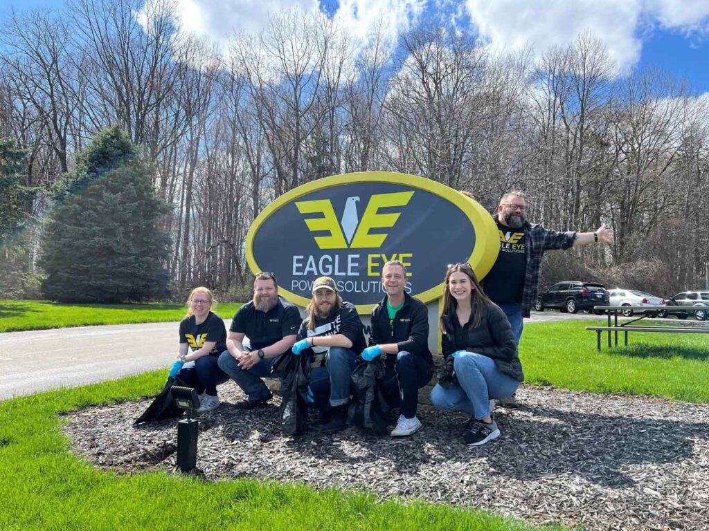 Team members of Eagle Eye Power Solutions proudly pose in front of their company sign after a successful outdoor cleanup effort, showcasing their commitment to community and environmental stewardship.