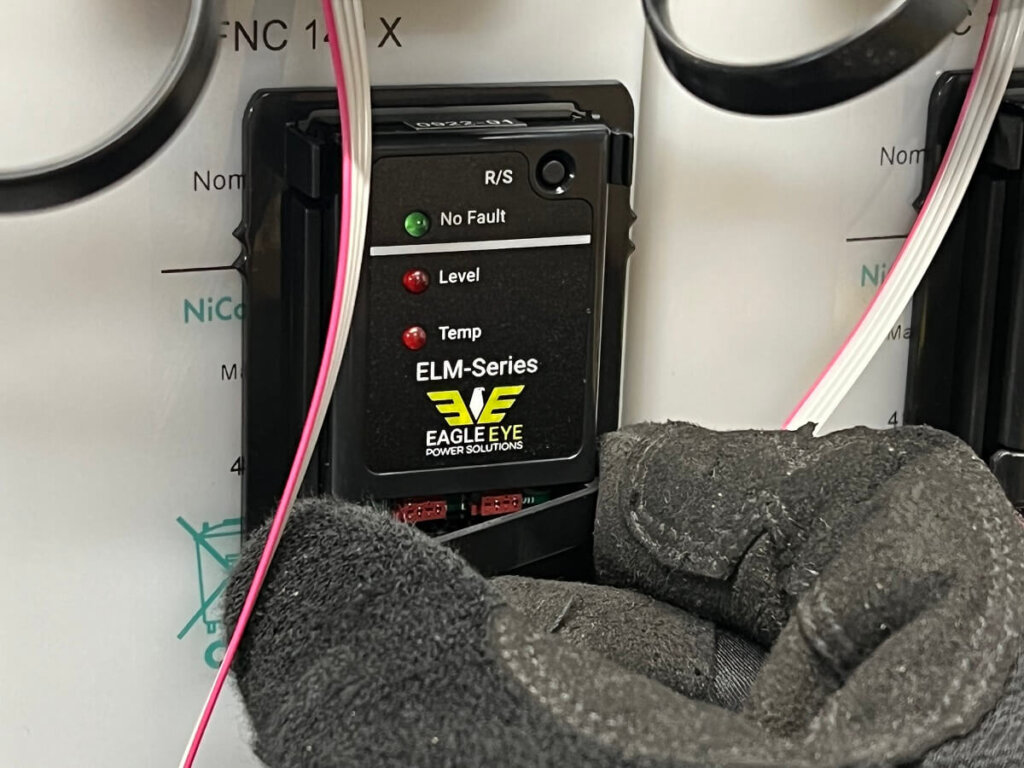 A gloved hand presses an Eagle Eye Power Solutions ELM-Series sensor into its mounted cradle on a battery jar. The sensor has indicator LEDs labeled “No Fault,” “Level,” and “Temp.” Pink and white wires are connected to the device.