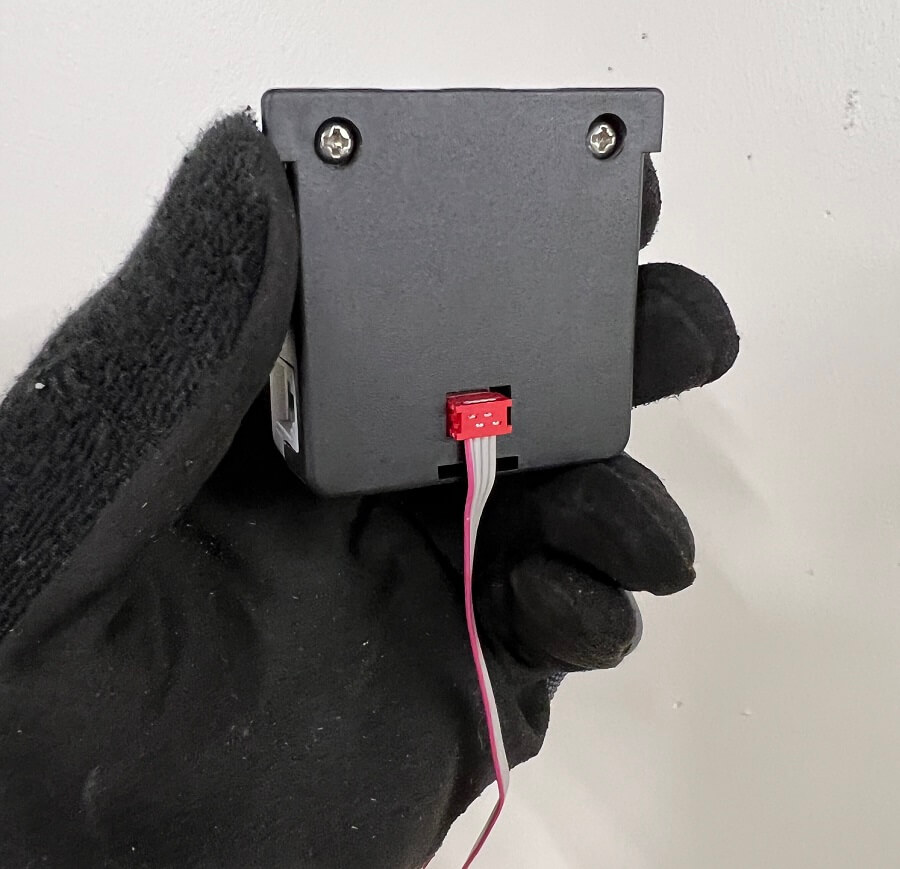 A gloved hand holds the back of an Eagle Eye Power Solutions Vigilant® battery sensor, showing a red ribbon cable securely connected to the rear ELM port. This connection is required for integrating the Electrolyte Level Monitor (ELM) as part of the installation process.