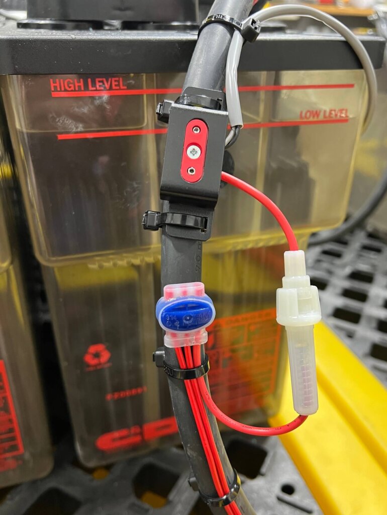 Close-up image of a battery charger cable with a red and black sensor clamp securely fastened to the cable using zip ties. Additional red wiring is zip-tied and organized neatly along the cable, with a visible blue splice connector and a white fuse holder hanging openly for later installation. The background shows a flooded-style battery with "High Level" and "Low Level" electrolyte markers.
