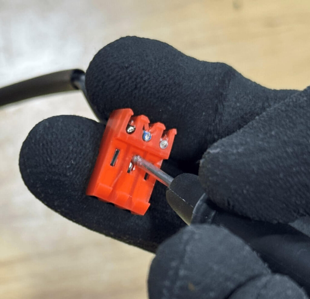 A person wearing black gloves holds a red IDC connector while a multimeter probe touches the exposed front pin to measure resistance. The connector has three visible metal contacts and is shown in close-up, demonstrating where to place the test lead during verification.