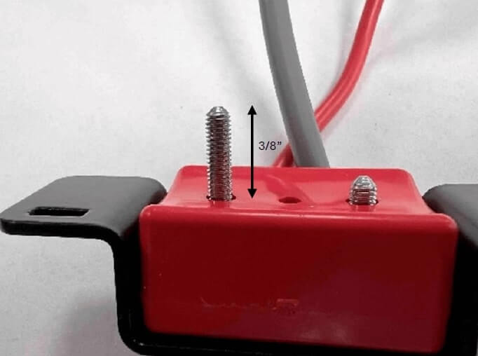 Close-up image of a red clamp with two metal grub screws partially inserted. A black double-headed arrow with a label "3/8”" shows the maximum penetration depth from the base of the mold into the cable. Red and gray wires are connected to the clamp in the background. This image demonstrates the correct screw depth when installing a clamp on a battery cable.