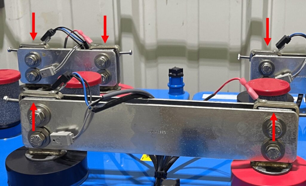 Four battery terminal clamps are shown installed on battery posts, each marked with a red arrow pointing to the adjustment screw. Cables are connected to each clamp, and care has been taken to ensure the clamps do not contact the bus bar. The clamps are uniformly oriented for consistency and accessibility.