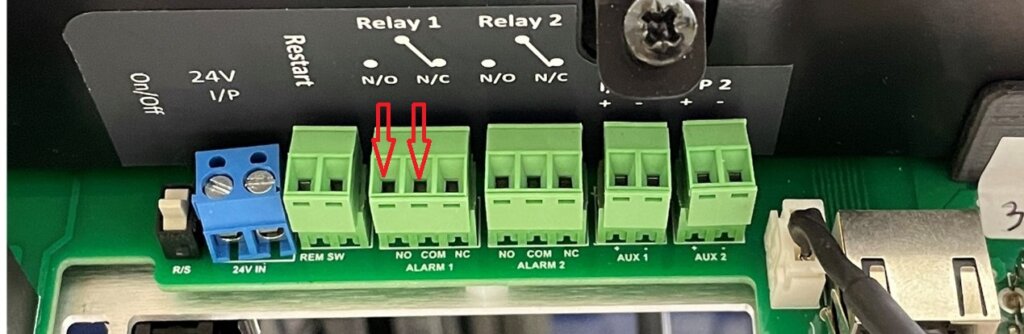 Close-up image of a circuit board showing green terminal blocks labeled for Relay 1, Relay 2, AUX 1, and AUX 2 on an Eagle Eye Power Solutions monitor. Red arrows highlight the "NO" (Normally Open) and "C" (Common) terminals of Relay 1, which are used to wire in a critical battery alarm. Labels indicate relay wiring points, and the image supports instruction for wiring Relay 1 to trigger on critical battery faults.