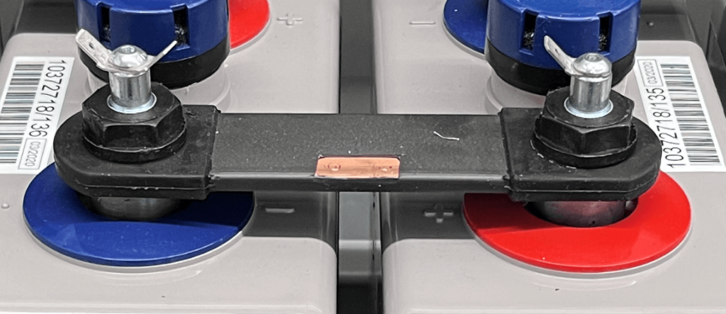 Close-up image of two battery terminals connected by a black intercell connector with a copper strip exposed in the center. Each terminal has a standoff bolt with wing nuts securing the connection. The left battery post is blue (negative), and the right is red (positive), following standard polarity markings. No clamps are used, indicating a standoff bolt-only configuration.
