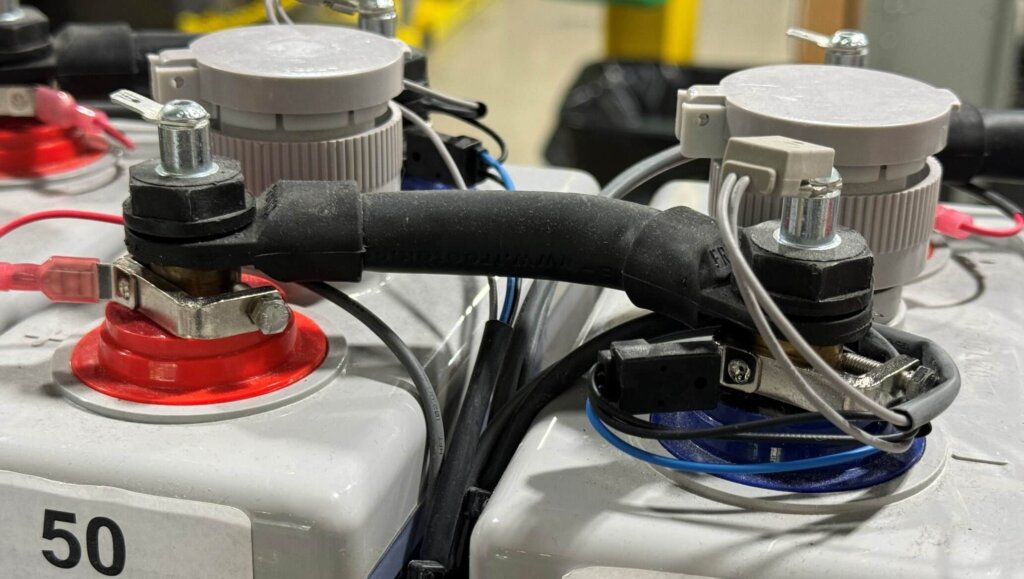 Close-up of battery terminals showing a standoff bolt installation with post clamps on both the positive and negative posts. The red terminal features a red sense wire and clamp, while the blue terminal includes a standoff bolt with gray and black sense and pulse leads. Both terminals are equipped with tab washers for proper electrical connections, demonstrating a typical configuration for Vigilant® battery monitoring systems.