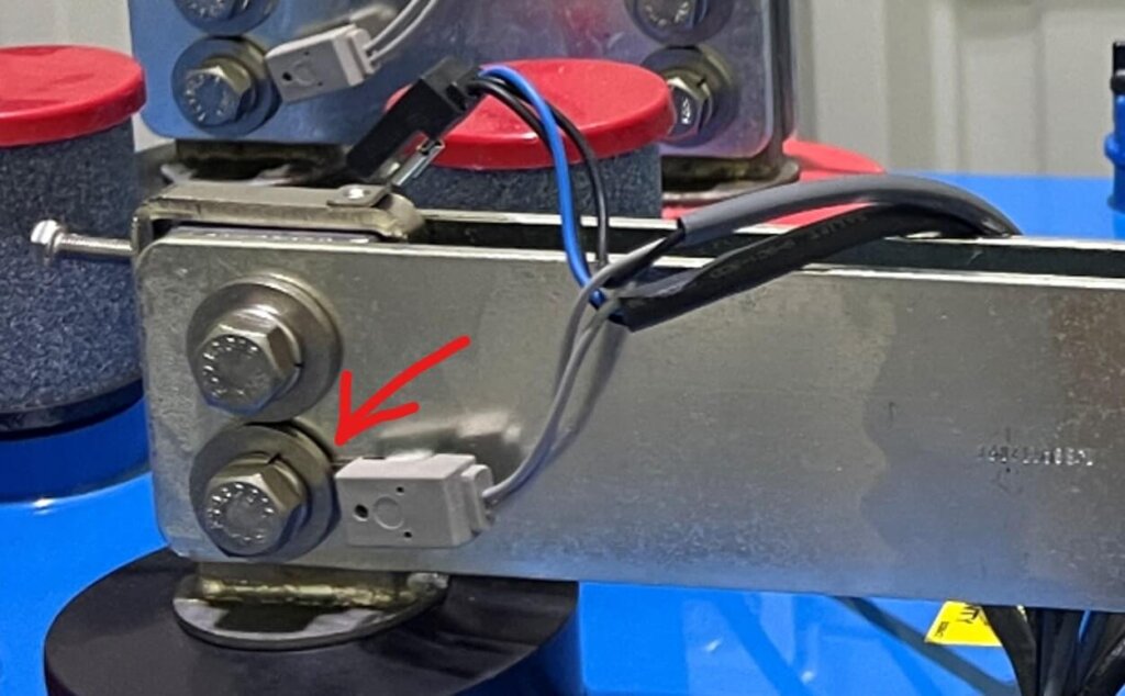 Close-up image of a battery terminal setup showing a gray tab washer installed on the bolt head side of the negative post clamp. A red arrow points to the tab washer, emphasizing proper installation placement. The clamp is securely fastened with two bolts, and sensor wires are attached, facing toward the Vigilant® monitoring system.
