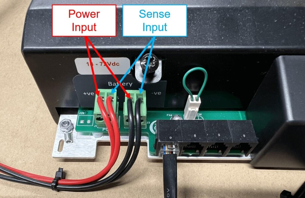 Close-up of the Vigilant® monitor's connection terminals showing power and sense inputs. Two red and two black wires are connected to the left Molex connector labeled "Power Input," while another set of black wires is connected to the adjacent Molex labeled "Sense Input." Labels and arrows indicate where each wire should be connected, with the power input on the left and the sense input on the right. A green jumper wire is connected beside the sense input block.