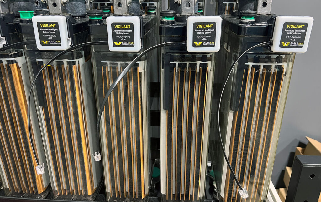 A row of transparent battery cells equipped with Eagle Eye Power Solutions' Vigilant® advanced intelligent battery sensors, each securely mounted in its cradle. Black communication cables and ELM (electrolyte level monitor) cables are connected to the sensors, indicating full installation and proper connectivity across the battery bank.