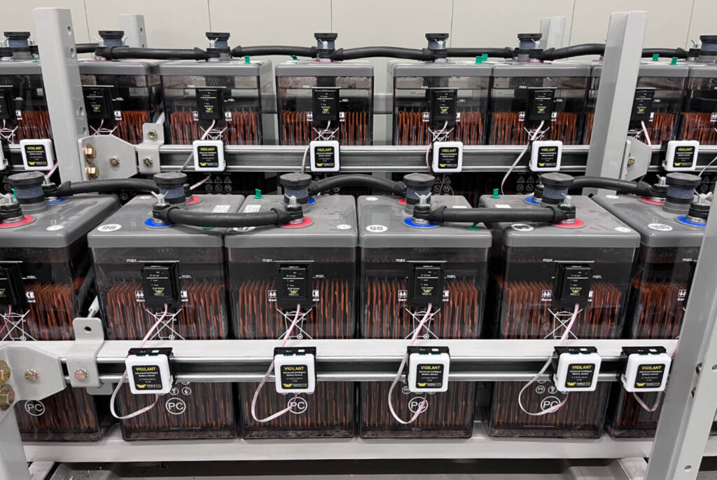 A battery string setup with multiple transparent battery jars, each equipped with Vigilant® sensors mounted on the front and ELM-Series sensors attached to the battery tops. White ribbon cables connect the ELM sensors to the Vigilant® sensors. The sensors are secured to mounting rails and all covers are in place, indicating completed installation.
