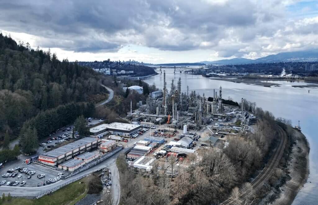 Aerial view of Tacoma Power facility beside a river, with industrial infrastructure, forested hills, and a bridge visible in the background.
