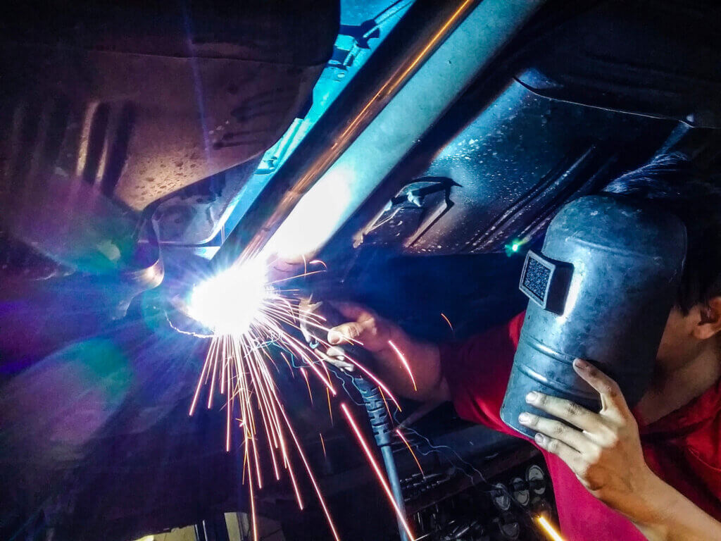 Blue-collar technician welding a car’s undercarriage in a garage, wearing gloves and eye protection as bright sparks fly from the torch.
