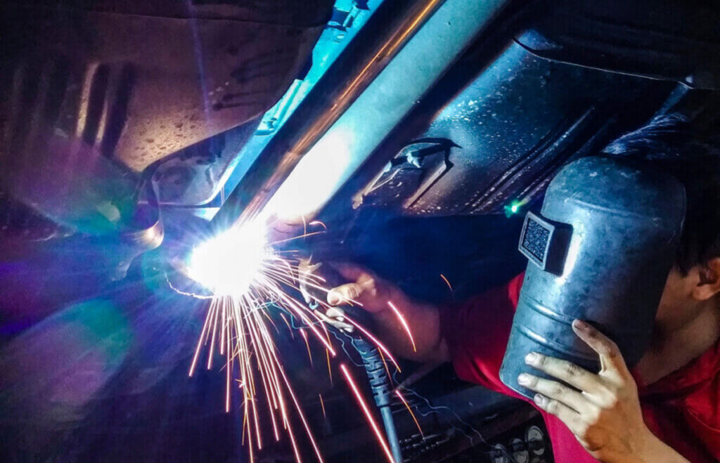 Blue-collar technician welding a car’s undercarriage in a garage, wearing gloves and eye protection as bright sparks fly from the torch.