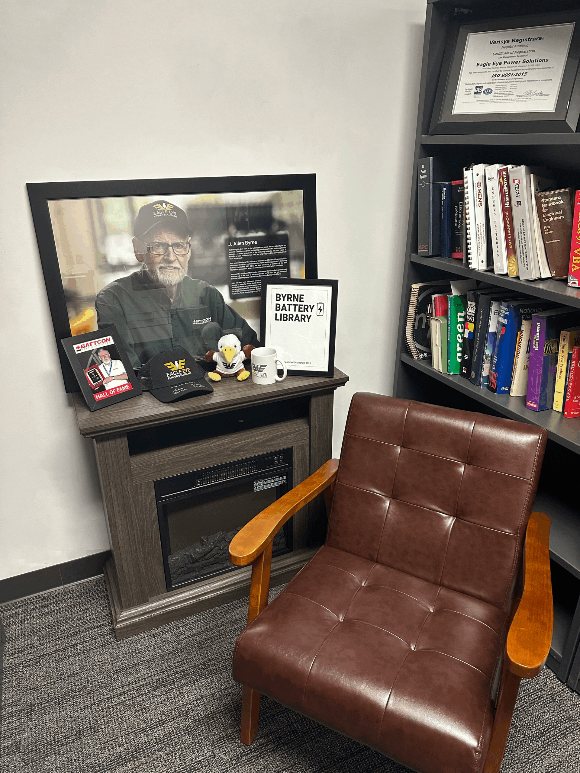 Cozy Byrne Library corner at Eagle Eye Power Solutions headquarters in Mequon, WI, featuring Allen Byrne’s portrait above an electric fireplace, Eagle Eye memorabilia, and a leather reading chair beside shelves of technical battery and power-system books.